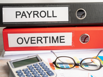 photo of binders labeled PAYROLL and OVERTIME on desk near calculator and eyeglasses 