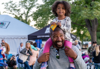 smiling black man in busy park carrying a young girl on his shoulders